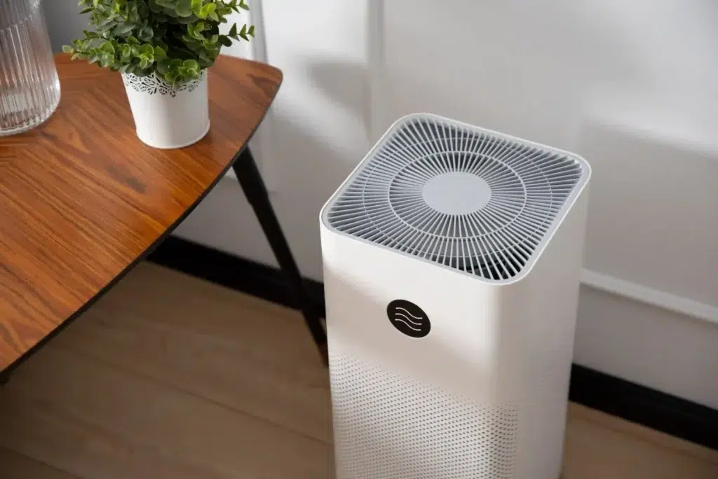 A white air purifier sits beside a wooden table, with a small potted plant and a glass pitcher, enhancing a tidy indoor space.