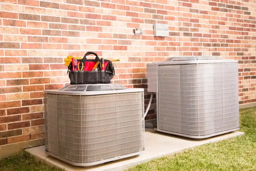Two outdoor HVAC units sit beside a brick wall, with a toolbox on top of one unit.