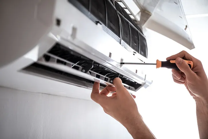 Hands using screwdriver to service wall-mounted air conditioner with open front panel and exposed components