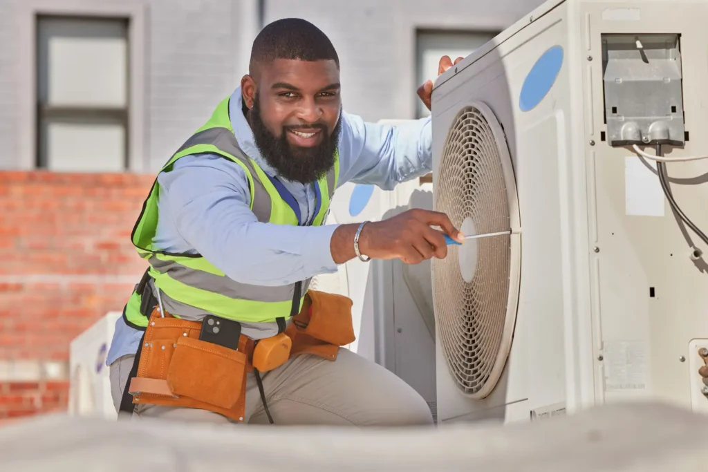 A worker in a green safety vest is inspecting an air conditioning unit, using a tool to tighten or adjust parts.