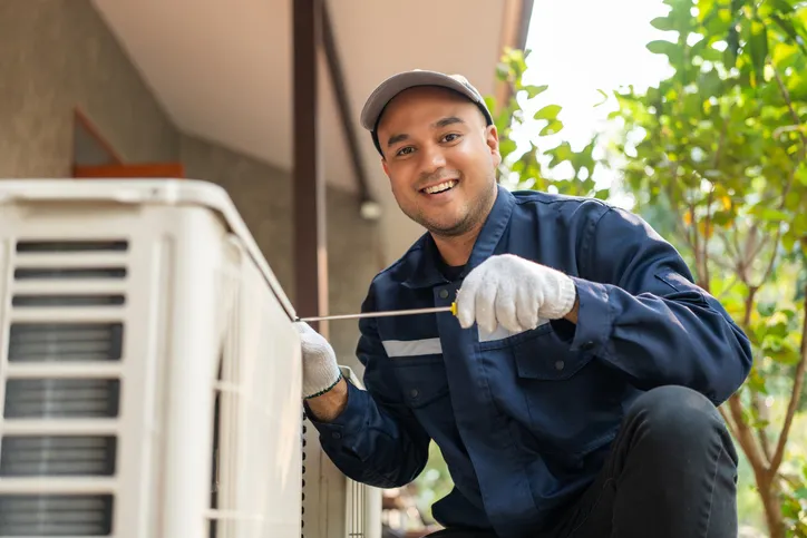 A smiling man wearing a cap, gloves, and a blue work uniform is crouched down, using a screwdriver on what appears to be an outdoor HVAC unit.