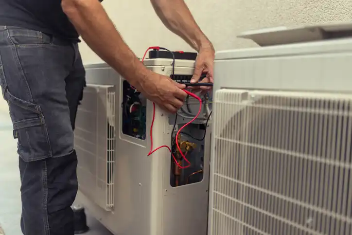 A technician services an outdoor HVAC unit, possibly a heat pump, checking its electrical connections with a multimeter.