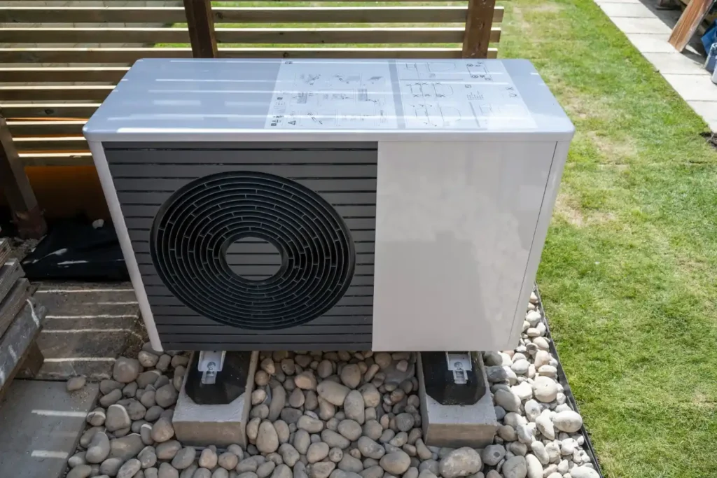 A large, white heat pump with a circular fan is placed on concrete blocks on a bed of gray stones next to a green lawn.