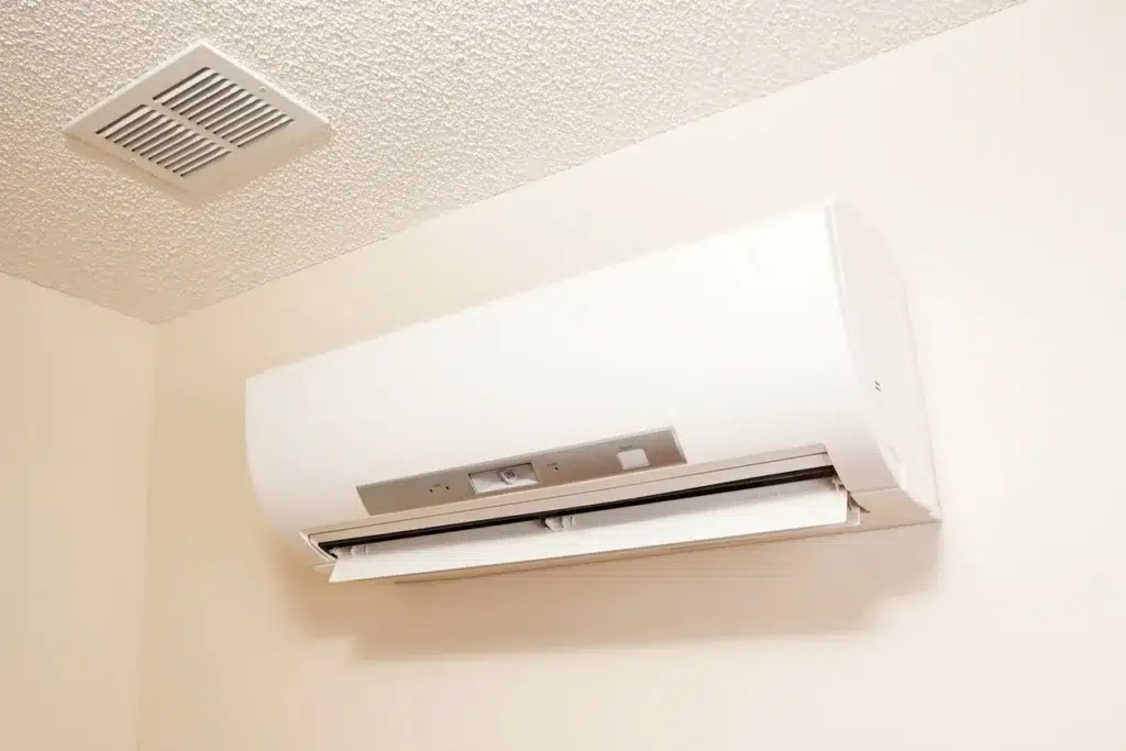 Wall-mounted white air conditioning unit with open vent in room corner near ceiling vent