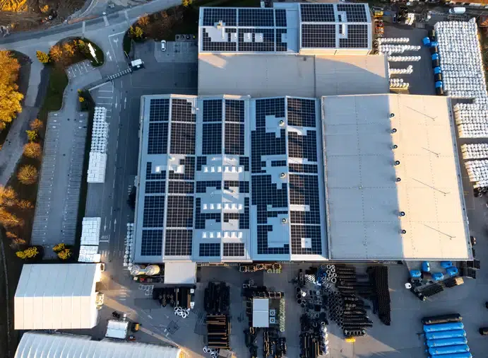 Aerial view of an industrial facility featuring solar panels on the roof, surrounded by stacks of materials and organized parking.