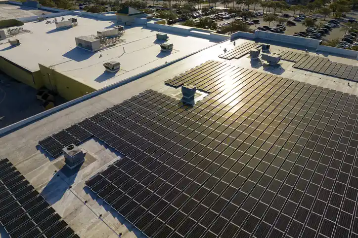 Aerial view of a commercial rooftop covered in solar panels, reflecting sunlight, with ventilation units and a busy parking lot nearby.