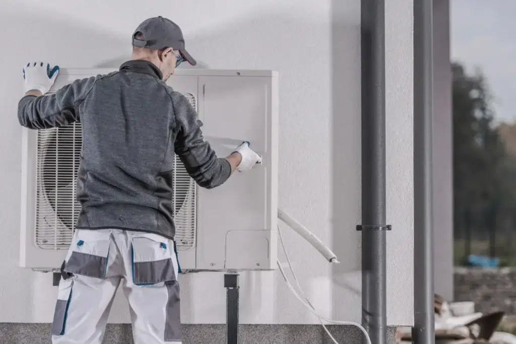 A technician in grey sweater and cap is replacing a heat pump unit attached at the wall of a house